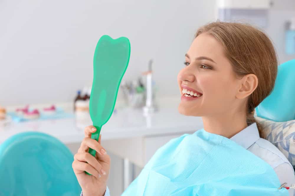 Young woman at Albanian dental clinic holding a mirror to check her smile after dental treatment.